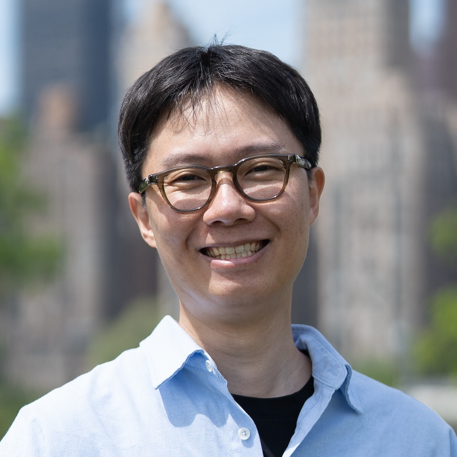 Photograph of a Asian man with short brown hair, eyeglasses, an blue shirt, standing in front of buildings and trees.
